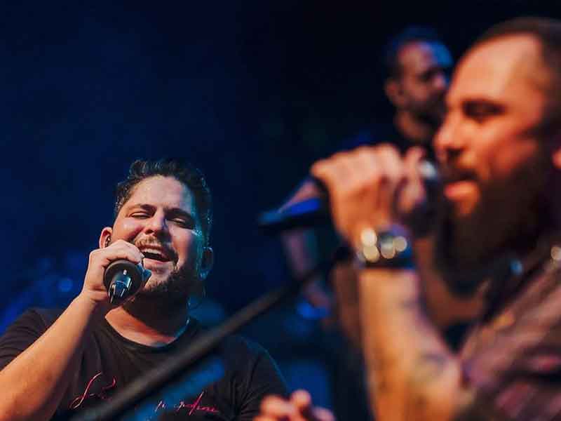 Foto durante show, Jorge à esquerda com microfone na mão vestido com camiseta preta. Ao lado, Mateus, cantando, segurando um microfone no pedestal. Mateus usa barba longa.
