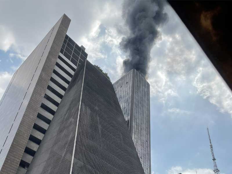 Na foto, aparece prédio com o topo emitindo grande quantidade de fumaça preta. Ao lado, prédio da FIESP, na Avenida Paulista em São Paulo.