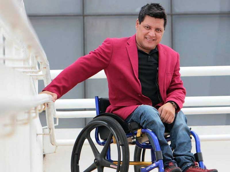 Wellington Camargo está em sua cadeira de rodas, vestindo blazer azul e calça jeans